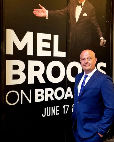 Todd Peterson standing next to a Mel Brook poster at a Mel Brooks related event