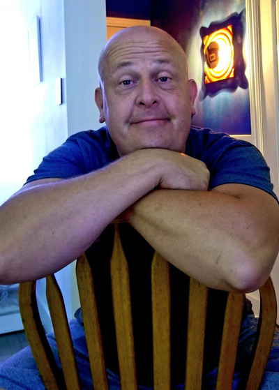 Smiling Todd Peterson sitting backwards on a wicker chair, arms folded along the top arch of the chair.